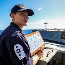 blue uniformed leading Steward wearing blue baseball cap looking to right whilst checking course notes on board with blue sky in background and another vessel alongside