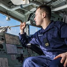side view of blue uniformed brown short haired Able Seaman on the bridge of HMS Sutherland Type 23 frigate looking out of window