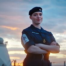 A Royal Navy crew member wearing a blue beret stands on the deck proudly looking at the camera