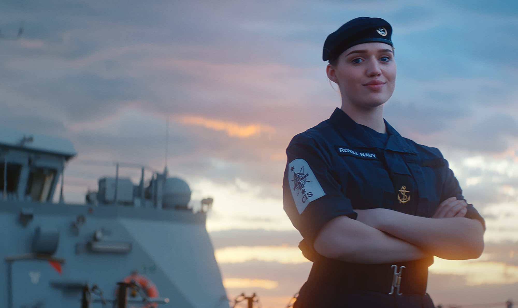 A Royal Navy crewmember wearing a blue beret stands on the deck proudly looking at the camera