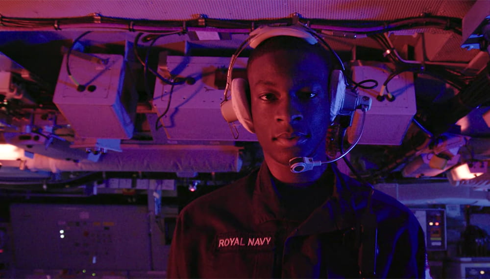 Royal Navy submariner wearing stands in a purple lit control room