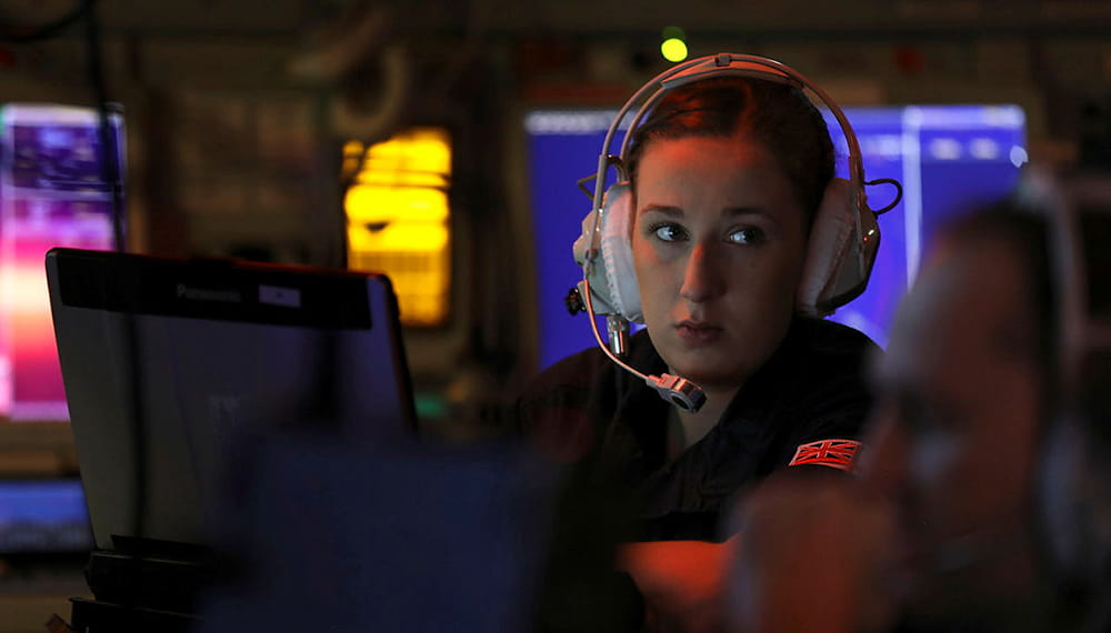 image in dark setting with monitors all around shows royal navy crewmember wearing headset looking sideways