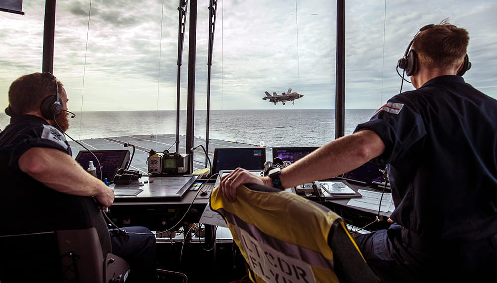with backs to the camera two uniformed royal navy sailors operating equipment in the flight control tower of aircraft carrier looking out over cloudy skies and grey sea