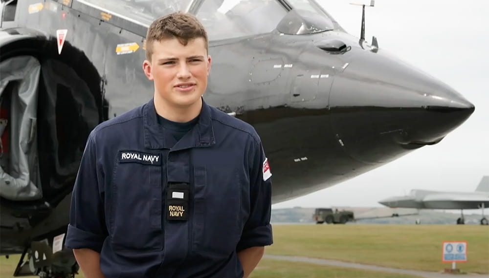 Royal Navy personnel looks towards camera with an aircraft in the background. 