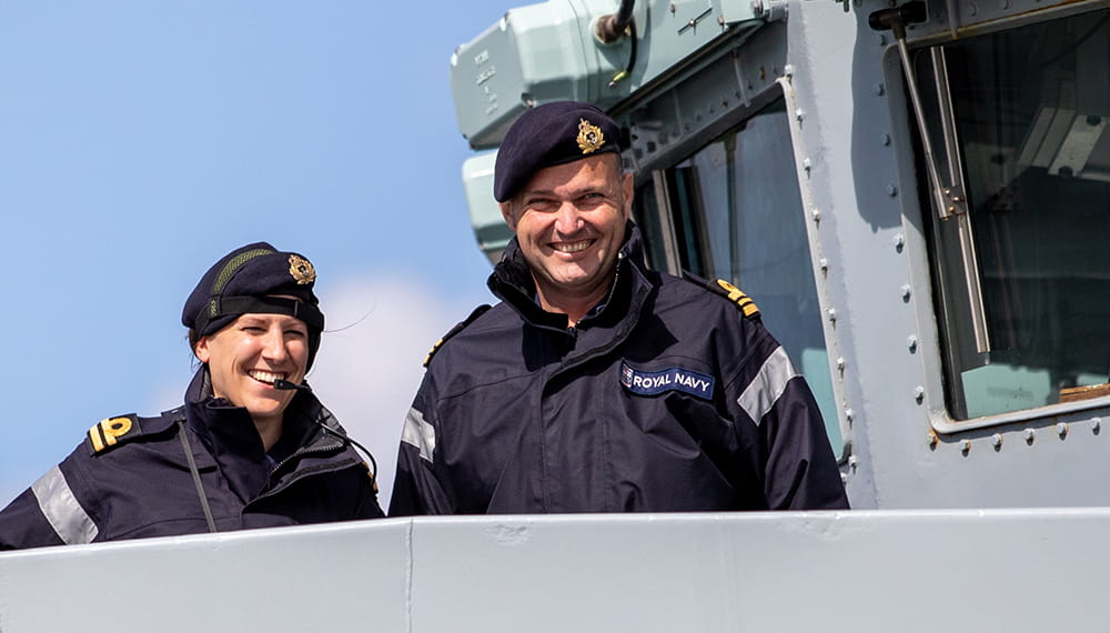 two smiling royal navy crewmembers in uniformed jackets on bridge of vessel smiling into camera in sunny conditions