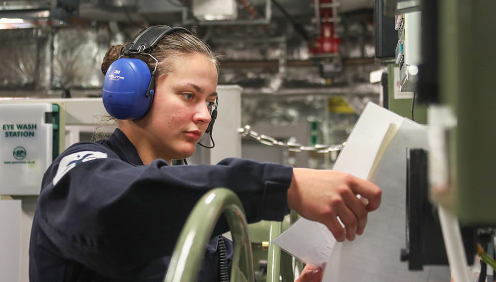 uniformed royal navy engineer wearing blue ear protectors looking at notes in engine room