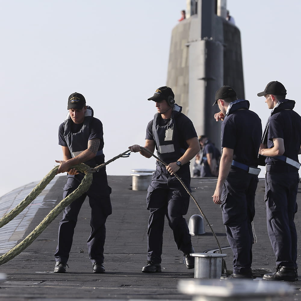four royal navy submariners in blue uniforms on deck of submarine pulling in ropes