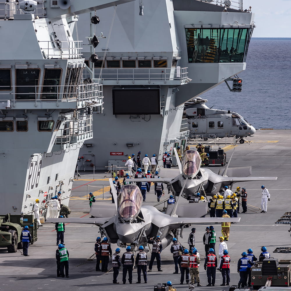 jets being prepared onboard aircraft carrier with control tower in background and aircraft and ground crew personnel on deck