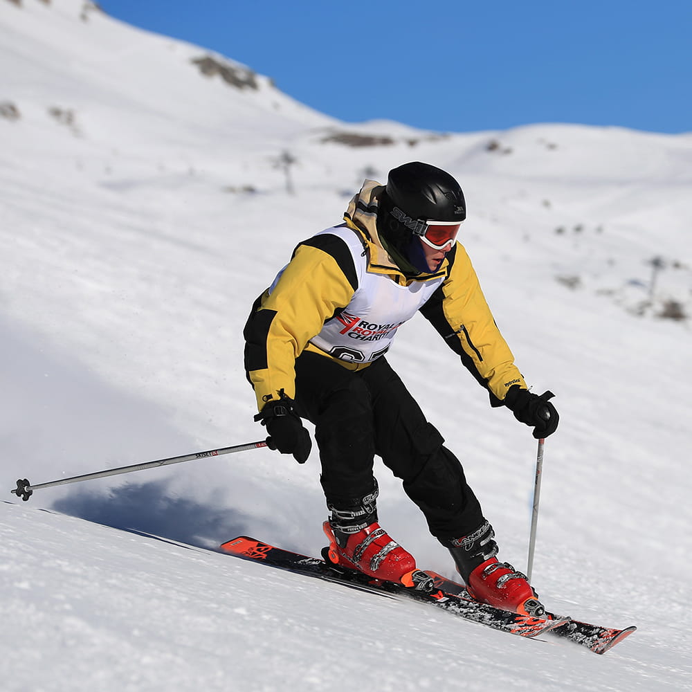 skier on slopes wearing blue trousers and yellow jacket with white racing vest with blue sky in background