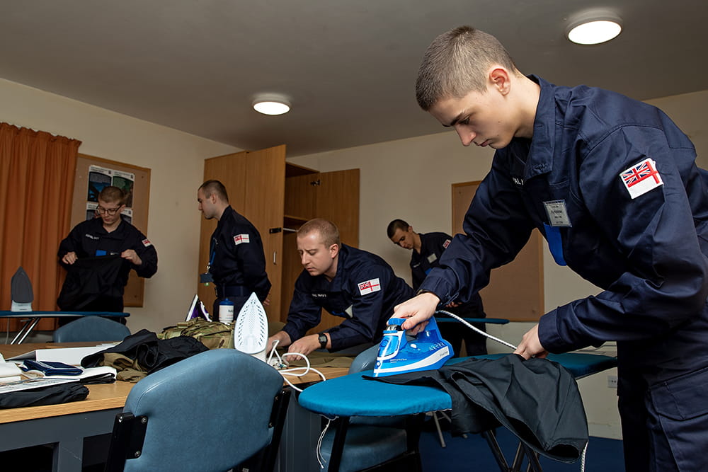 navy personnel in blue uniforms completing domestic tasks with rating in foreground ironing uniform