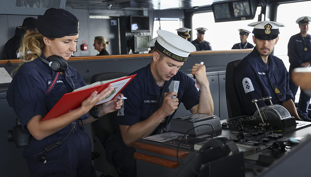 The ship's quartermaster on the bridge by the ship controls reads notes into a microphone as an officer and a rating watch