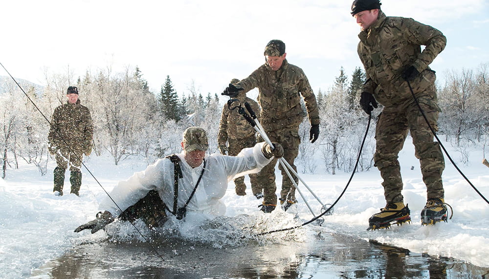 Royal Marine Reservist steps into icy water whilst other Reservists in cameo uniforms stand on the ice during training