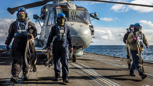 Aviation personnel walking along a flight deck with a helicopter in the background