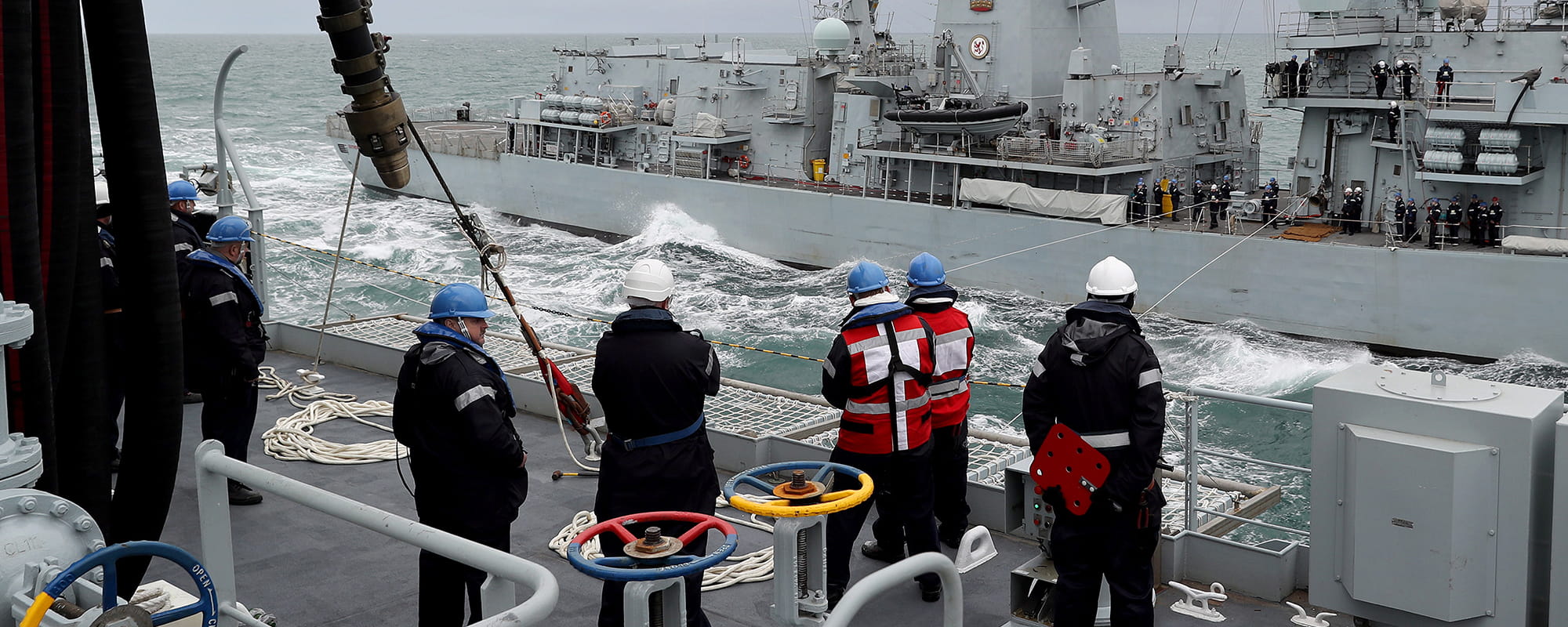 Crew members stand on deck looking at the opposing boat. RFA Tidespring conducts her first ever RAS with a Royal Navy Warship, HMS Sutherland. 