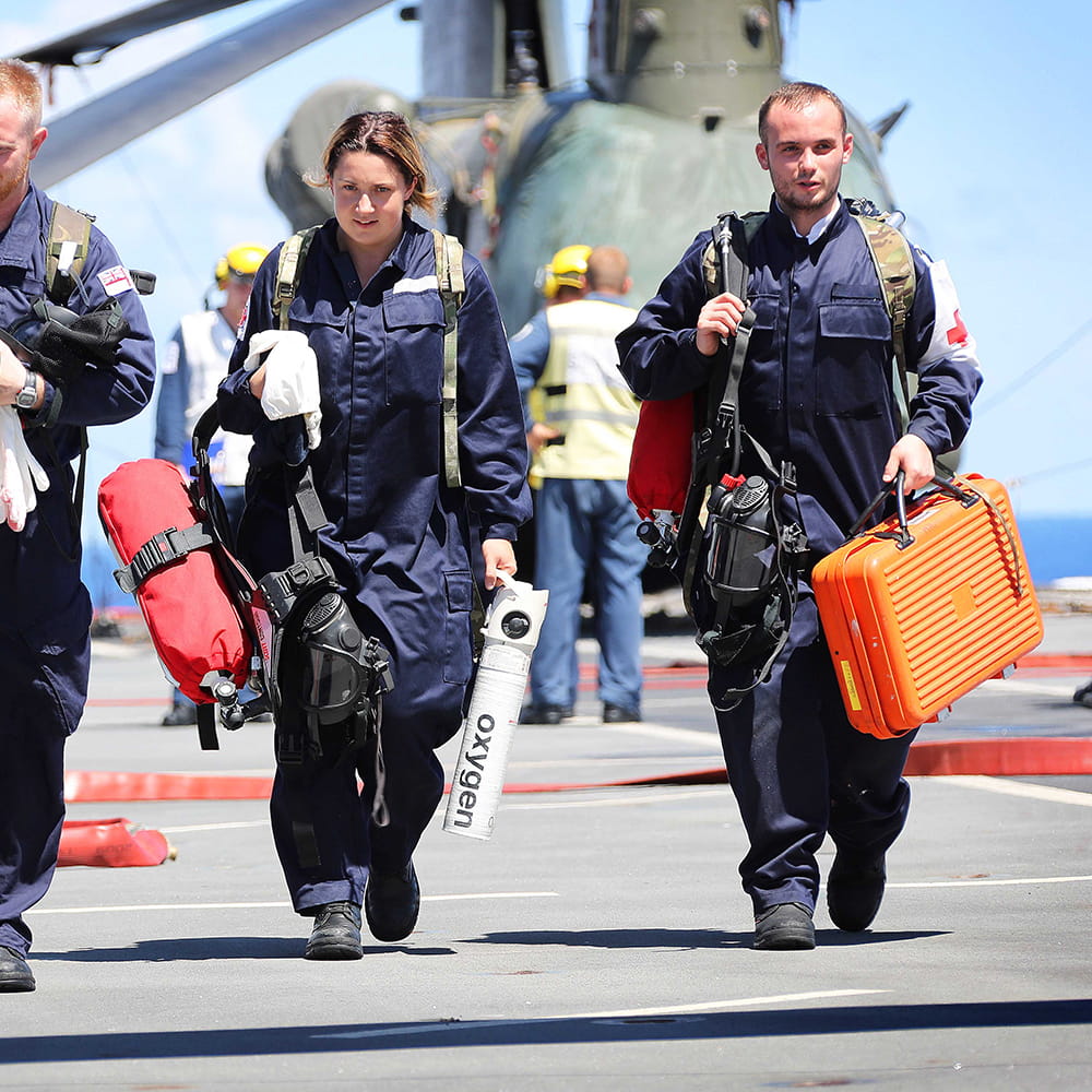 A man and a woman in navy overalls walk towards the camera carrying an oxygen tank, gas masks and an orange case