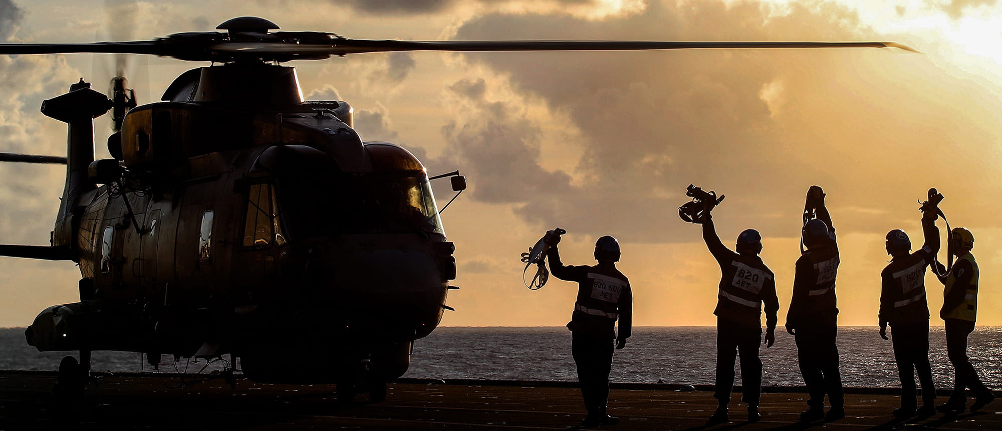 Royal Navy crewmembers waving at a helicopter on the flight at sunset