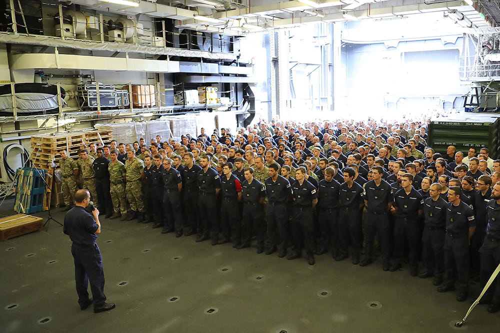 A large group of Royal Navy personnel stand together on a lower deck listening to a briefing