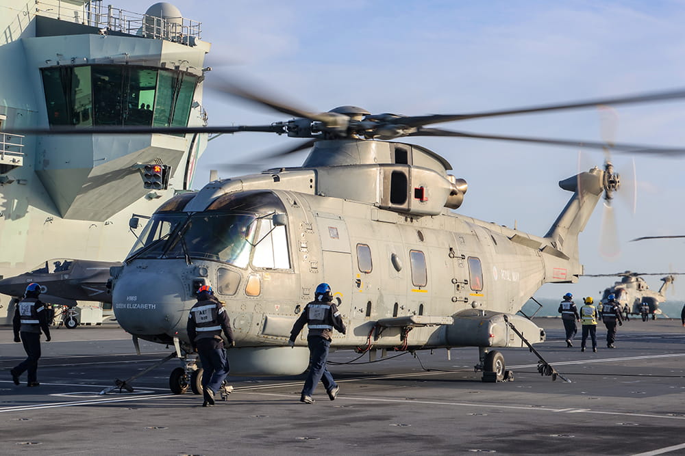 Royal Navy personnel walk around a Merlin helicopter onboard an aircraft carrier