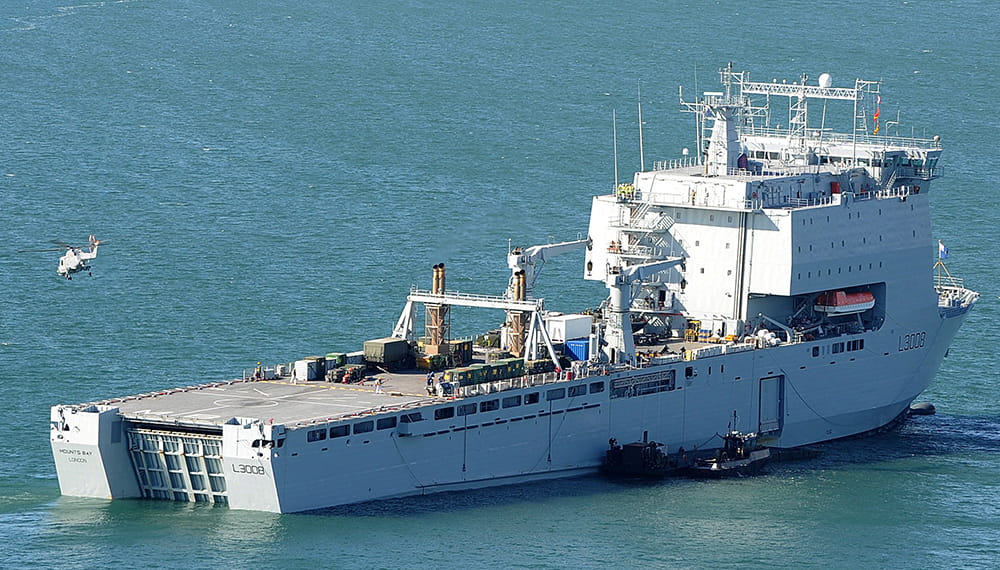 RFA Mounts Bay at sea, with a helicopter flying off into the distance
