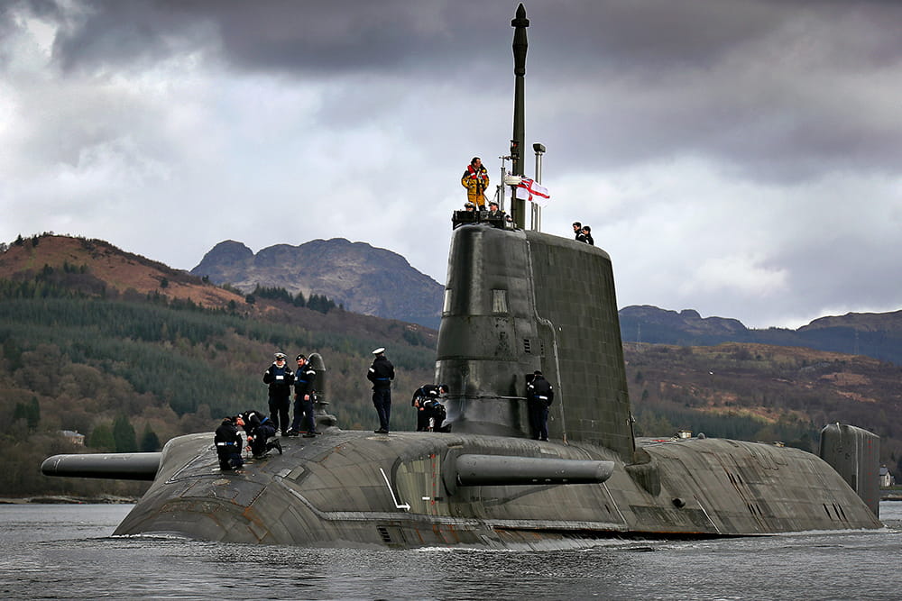 Several Royal Navy submariners stand on the on top of an Astute Class submarine