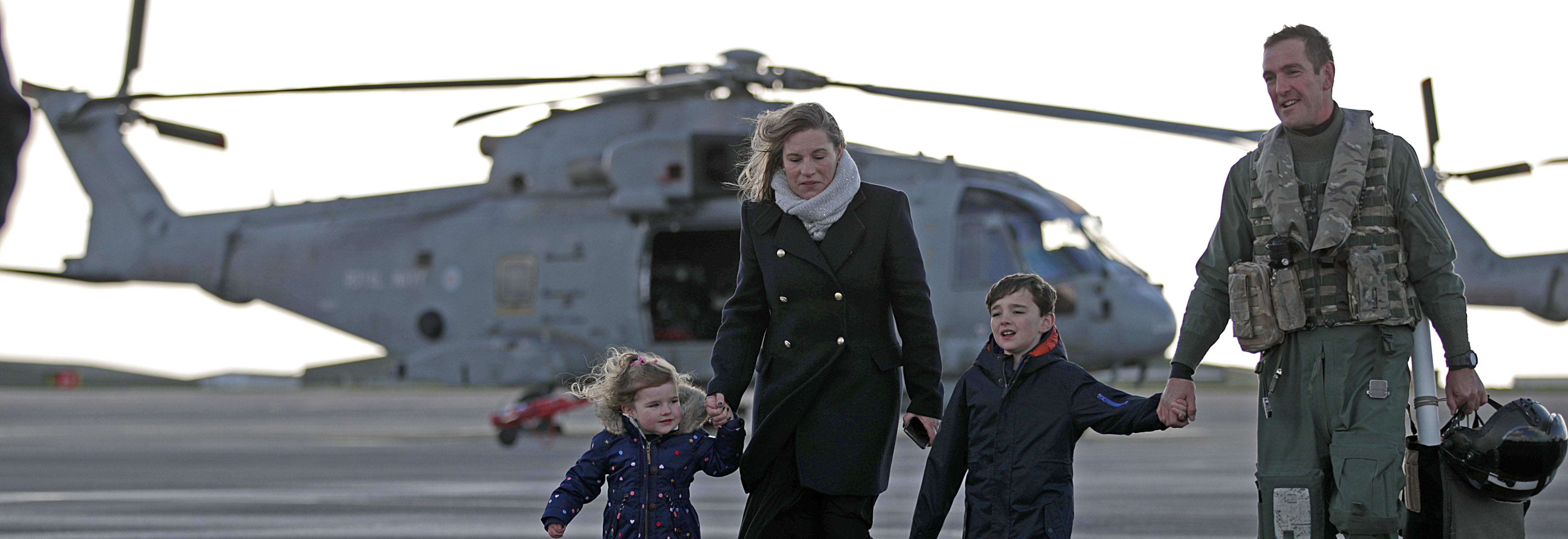 An airman and his family walks holding hands. In the background a Merlin helicopter.