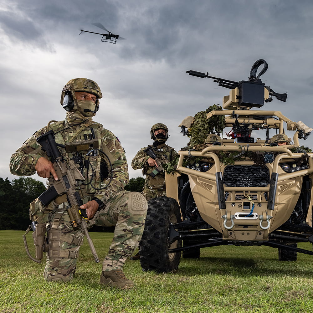 Royal Marine kneels beside a lightweight vehicle whilst a drone flies above