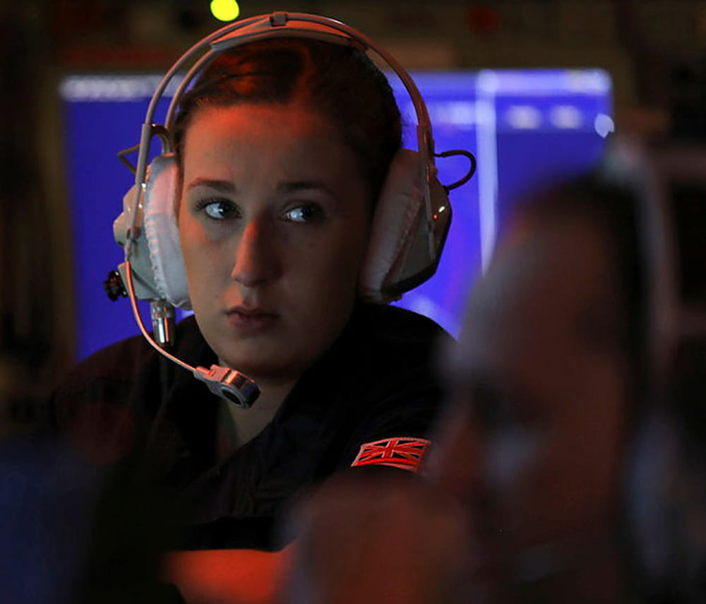 Image in a dark setting with monitors all around shows a Royal Navy crewmember wearing a headset looking sideways