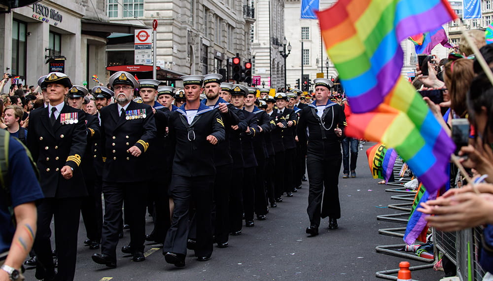 Royal Navy sailors march in front of crowd waving Rainbow flags