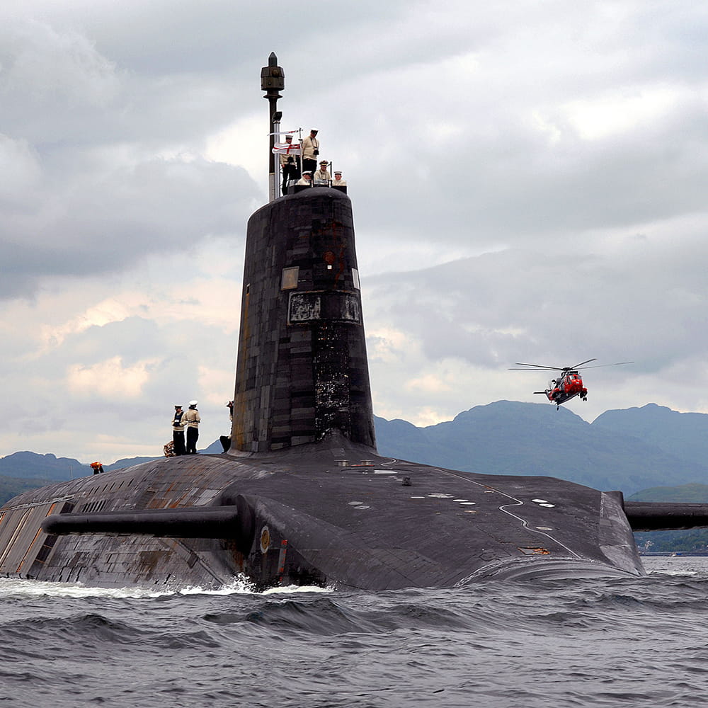 Royal Navy submarine on the surface with crew on the hull and helicopter flying overhead