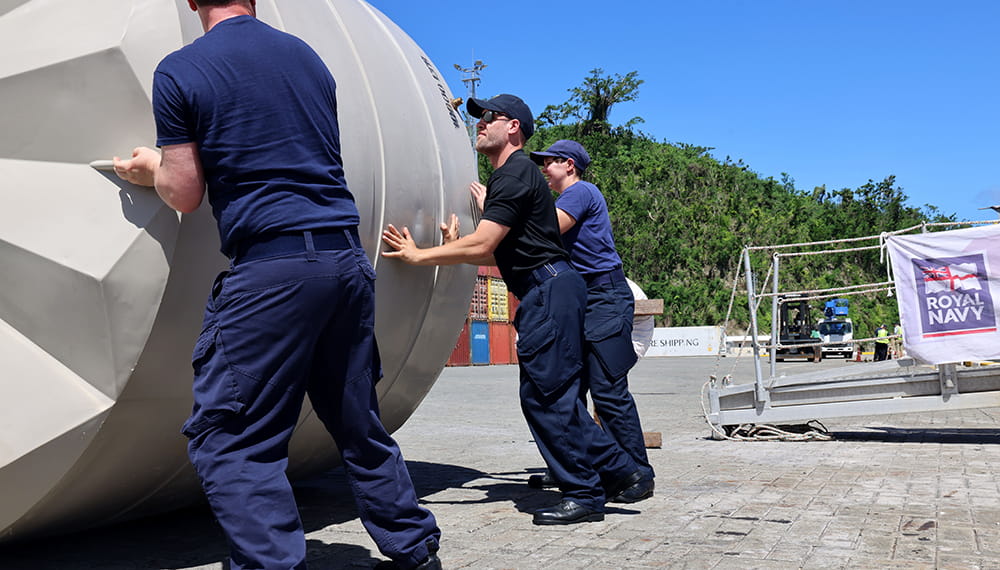 Three Royal Navy sailors role a huge humanitarian aid water tank along the ground